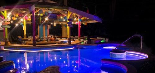 A vibrant poolside bar at night, illuminated with colorful lights and surrounded by a glowing blue pool.