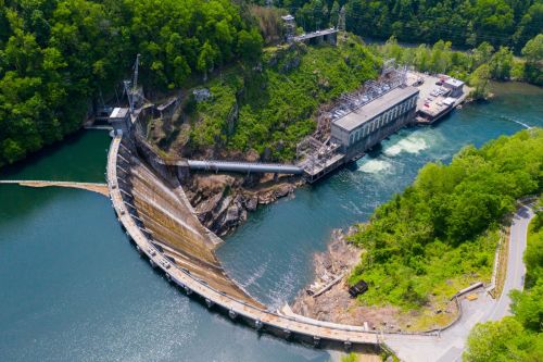 Aerial view of a dam with water flowing over it, surrounded by lush greenery and a power station nearby.