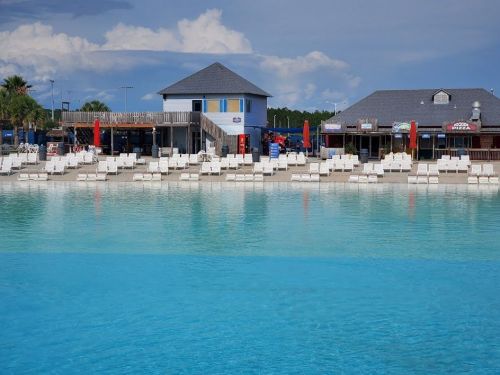 A serene pool area with white lounge chairs, a blue building, and clear turquoise water under a partly cloudy sky.
