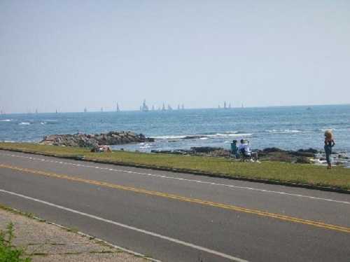 A coastal scene with a road, grassy area, and people enjoying the view of sailboats on the water in the distance.