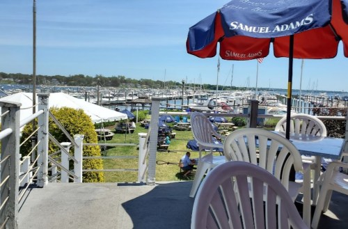 View from a patio overlooking a marina with boats, picnic tables, and a blue sky. A red umbrella is in the foreground.