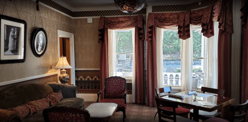 Cozy room with vintage decor, featuring a sofa, armchair, lamp, and large windows with curtains.