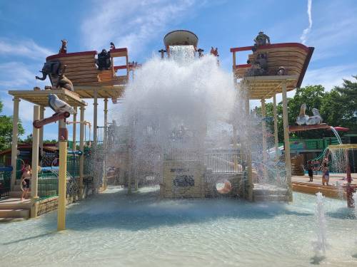 A water play area with a large splash feature, surrounded by children and colorful structures under a blue sky.