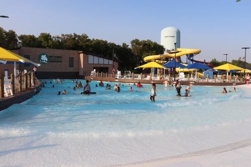 A vibrant water park scene with people enjoying a wave pool and water slides under sunny skies and colorful umbrellas.