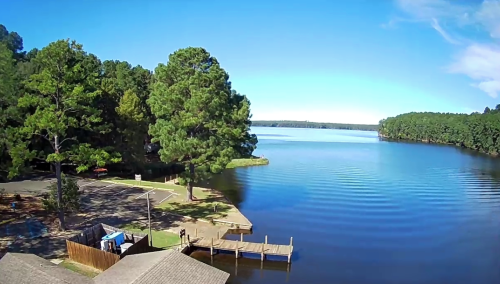 A serene lake view surrounded by trees, with a wooden dock and clear blue sky reflecting on the water.