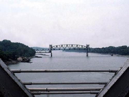 A view of a bridge spanning a river, surrounded by lush greenery and a cloudy sky.