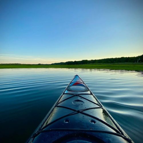 A kayak's bow cutting through calm water, with a clear blue sky and green shoreline in the background.