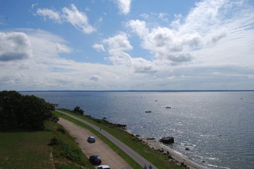 A serene coastal view with a winding road, cars, and boats on the water under a partly cloudy sky.
