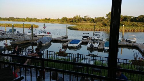 View of a marina with boats docked, surrounded by greenery and calm water, with people seated nearby.