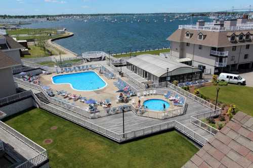 Aerial view of a pool area with sun loungers, surrounded by grass and a waterfront with boats in the background.