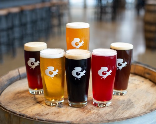 A variety of craft beers in glasses with a logo, arranged on a wooden barrel in a brewery setting.