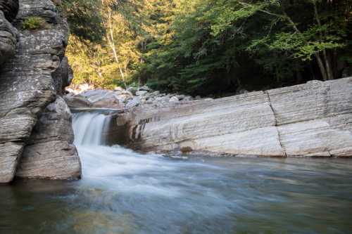 A serene waterfall cascades over smooth rocks into a tranquil stream, surrounded by lush greenery.