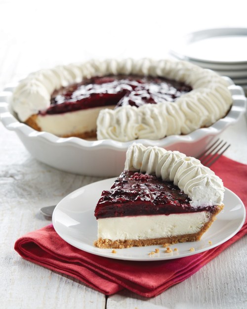A slice of cheesecake topped with berry glaze and whipped cream, served on a plate next to a whole pie.