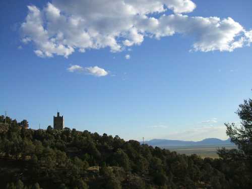 A scenic view of a tower on a hillside, surrounded by trees, with a blue sky and distant mountains in the background.