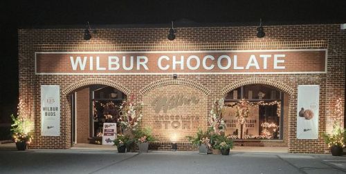 A brick storefront at night with "Wilbur Chocolate" sign, decorated with lights and plants.