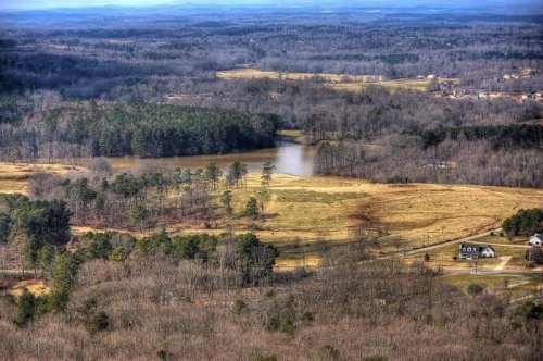 Aerial view of a serene landscape with a lake, fields, and scattered trees, under a cloudy sky.