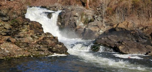 A cascading waterfall flows over rocky terrain, surrounded by trees and mist in a serene natural setting.