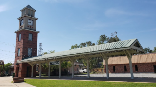 A clock tower beside a covered platform in a park-like setting, with blue skies and trees in the background.