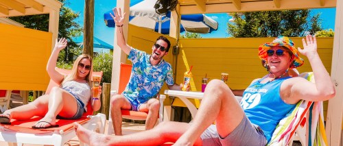 Three friends relax in colorful beach attire, waving and enjoying drinks in a sunny outdoor setting.