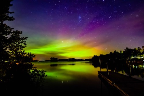 A serene lake at night, reflecting vibrant auroras and a starry sky, with silhouettes of trees and a dock in the foreground.