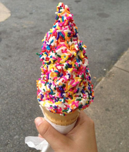 A hand holding a large ice cream cone topped with colorful sprinkles against a blurred outdoor background.