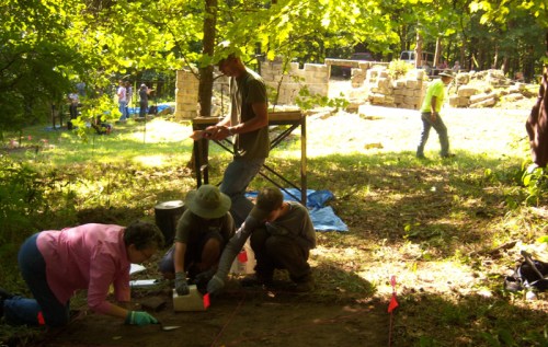 A group of people excavating at a site, with tools and flags marking areas, surrounded by trees and ruins in the background.