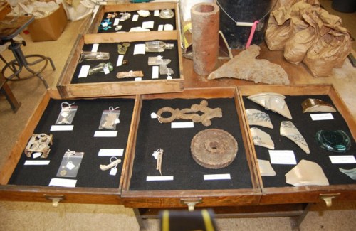 Display of various artifacts in wooden trays, including glass pieces, metal objects, and pottery shards.