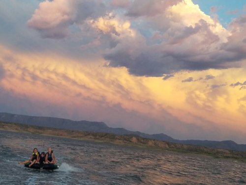 Two people enjoy tubing on a lake under a colorful sunset with dramatic clouds and mountains in the background.