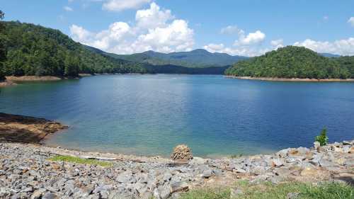 A serene lake surrounded by green hills and mountains under a blue sky with fluffy white clouds.