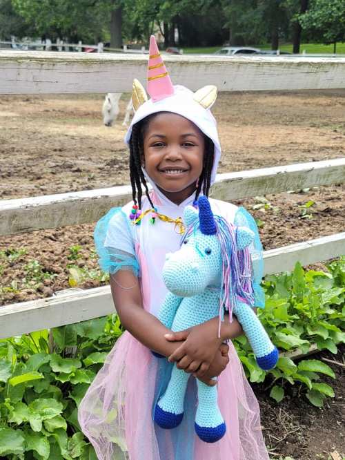 A smiling girl in a unicorn costume holds a blue crocheted unicorn toy, standing in a farm setting.