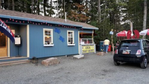 A blue building with a sign for local craft beers, surrounded by trees and outdoor seating, with a small car parked nearby.