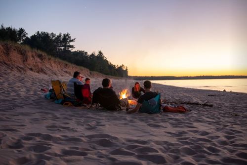 A group of people sitting around a campfire on the beach at sunset, enjoying a cozy gathering.