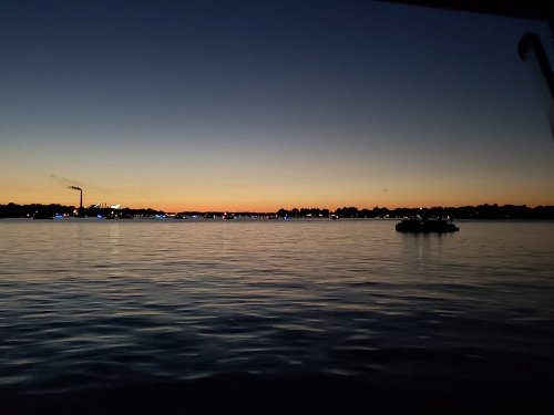 A serene sunset over a calm body of water, with silhouettes of trees and distant lights reflecting on the surface.