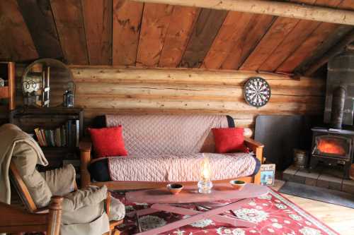 Cozy cabin interior with wooden walls, a sofa, a dartboard, and a wood stove, creating a warm, inviting atmosphere.