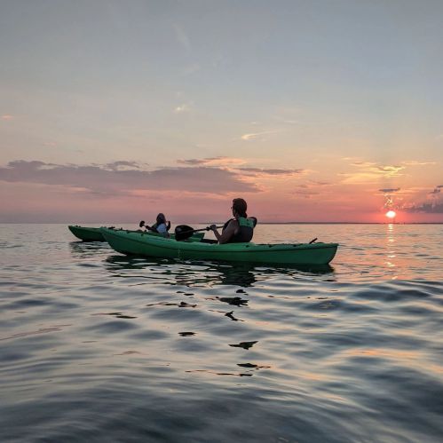 Two people kayaking on calm water at sunset, with vibrant colors reflecting on the surface.