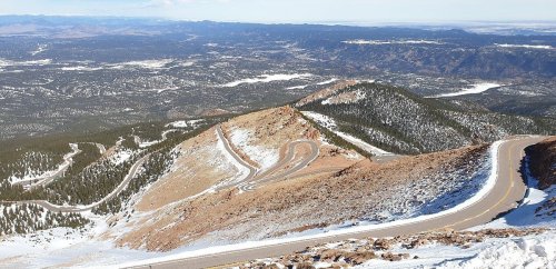 A winding road descends a snowy mountain, surrounded by a vast landscape of trees and distant hills.
