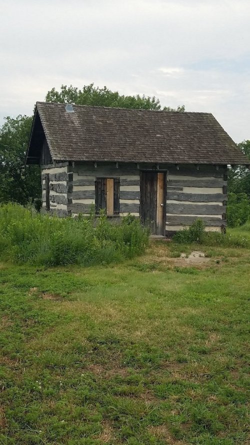 A small, rustic log cabin with boarded windows, surrounded by tall grass and trees in the background.