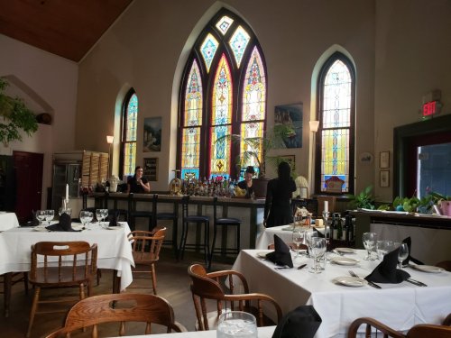 Interior of a restaurant with stained glass windows, tables set with white linens, and a bar in the background.