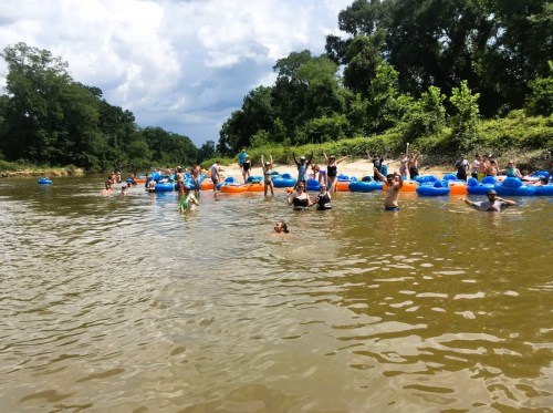 A group of people enjoying a river float, some in tubes, others swimming, surrounded by trees and a cloudy sky.