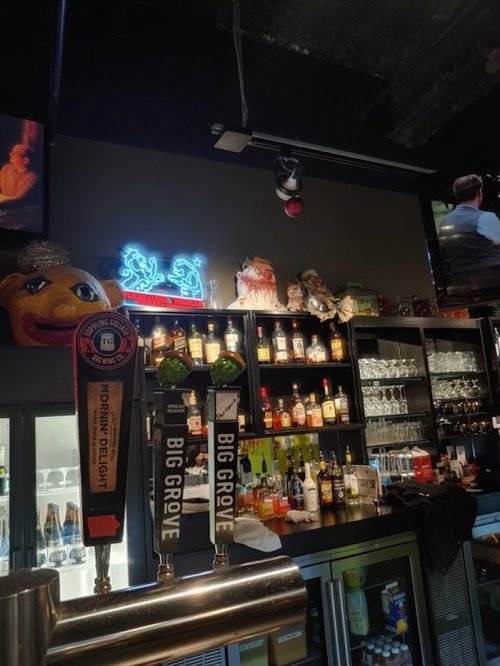 A bar scene featuring drink taps, shelves of liquor, and decorative items, with a neon sign in the background.