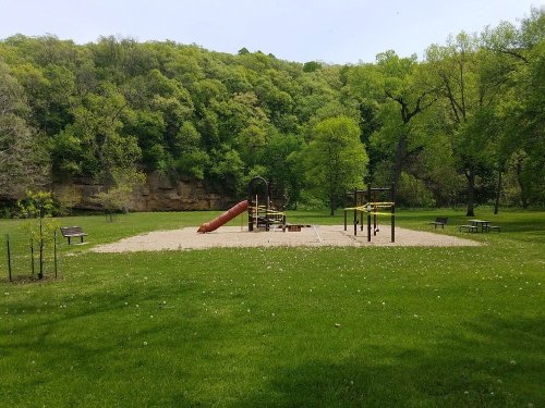 A playground with a slide and climbing structures, surrounded by green grass and trees in a park setting.