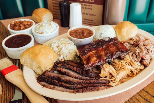 A platter of barbecue featuring brisket, ribs, pulled pork, coleslaw, baked beans, and rolls, with sauces on the side.