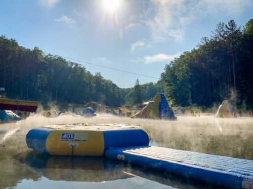 A misty lake scene with inflatable water obstacles and trees in the background under a bright sun.