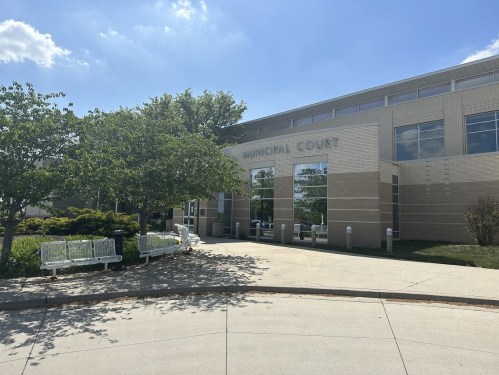 Exterior view of a modern municipal court building with trees and benches in front, under a partly cloudy sky.