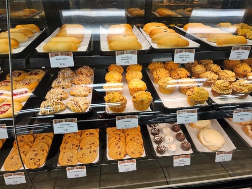 A display case filled with various baked goods, including cookies, muffins, and pastries, all labeled with prices.