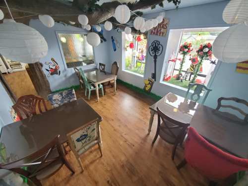 Cozy café interior with wooden floors, decorated tables, paper lanterns, and colorful floral arrangements in windows.