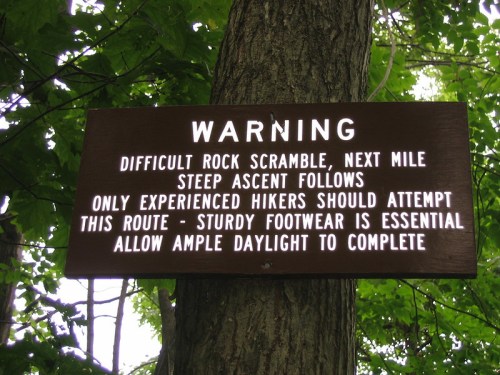 A warning sign on a tree about a challenging rock scramble ahead, advising experienced hikers to wear sturdy footwear.