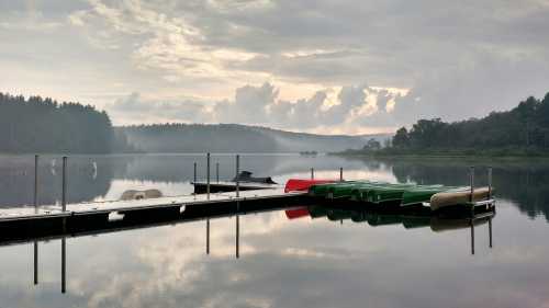 A calm lake at dawn with a wooden dock and colorful boats, surrounded by misty trees and soft clouds.