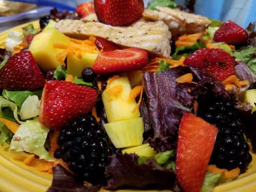 A colorful salad topped with grilled chicken, strawberries, blackberries, mango, and shredded carrots on a bed of greens.