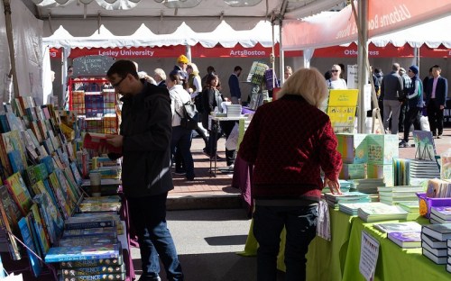 A busy outdoor book fair with people browsing tables filled with colorful books under a large tent.